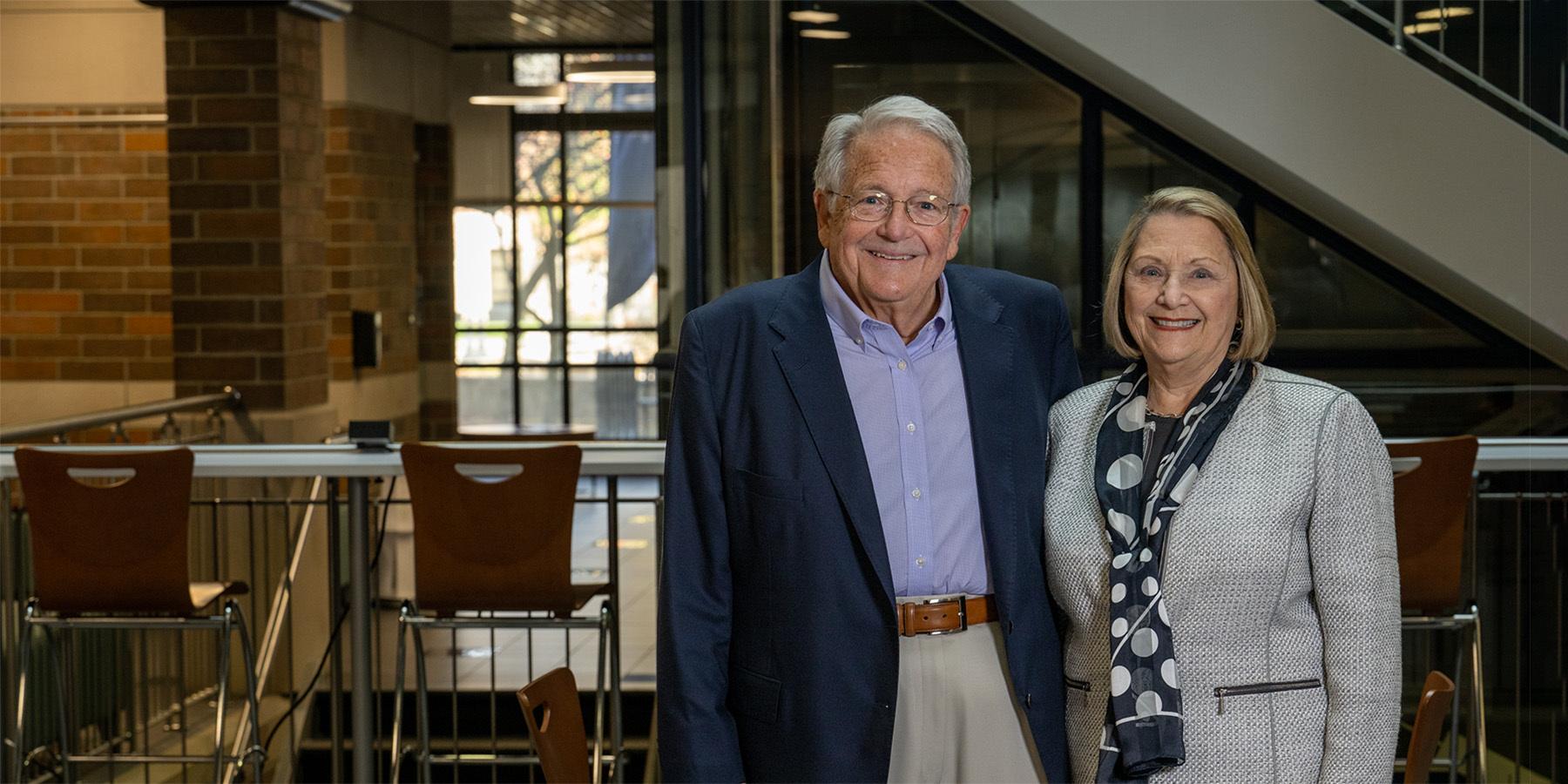 Gloria (left) and Steve (right) Bailey stand smiling in the Bailey College of Engineering & Technology Building. She is wearing a beige business casual suit with a black scarf that has large white circles on it. He is wearing a dark blue sport coat with light blue shirt, beige khakis, and brown belt. The visible interior of the building is dark, with outdoor sunlight visible through windows in the back of the building.